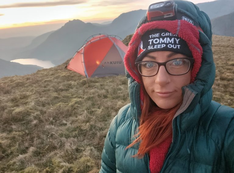 Woman camping on top of grassy hill with tent and lake in the background wearing The Great Tommy Sleep Out beanie hat