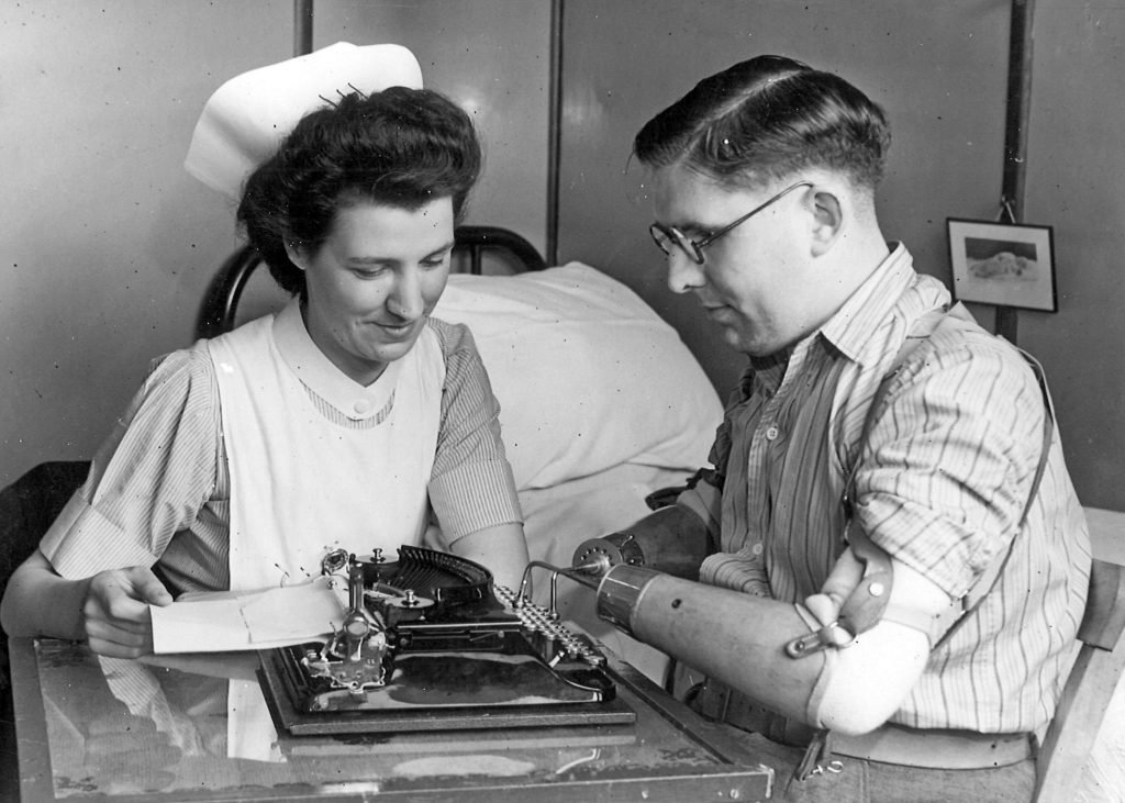 A black and white photo from 1919. A nurse teaches a disabled veteran, who lost both hands, to type using a typewriter with his new hook prosthetic.