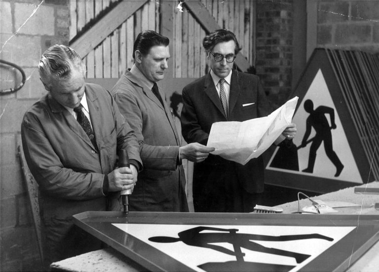 Employee creating a sign with colleagues looking at plans in the background