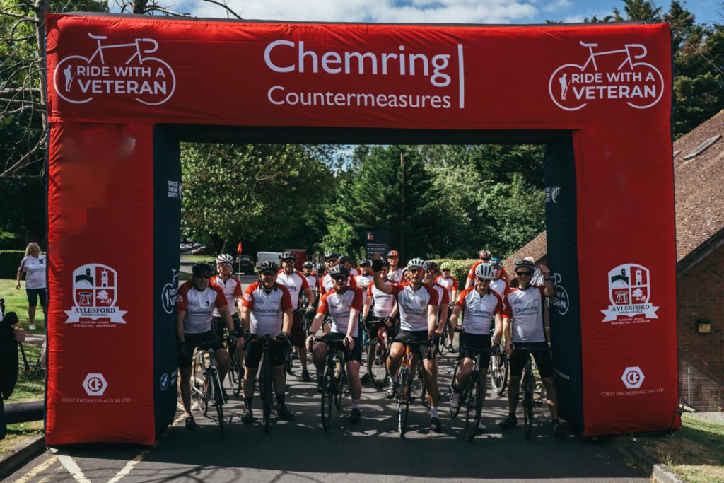 Cyclists lining up in their gear and on their bikes beneath a Ride with a Veteran sign